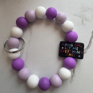 May include: A beaded keychain bracelet in a circular shape. The bracelet features a mix of white, lavender, and purple beads. A small black rectangular charm with the text "HANG ON, *LET ME OVERTHINK THIS*" is attached to the bracelet.