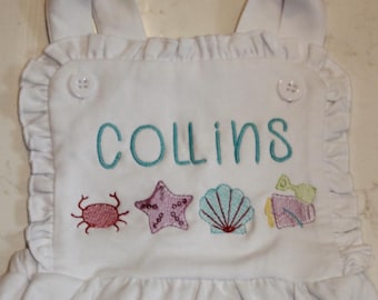 May include: White baby romper with ruffled edges and straps. The romper features the name "COLLINS" embroidered in teal, along with embroidered sea-themed designs including a crab, starfish, seashell, and a gift. Buttons secure the straps.