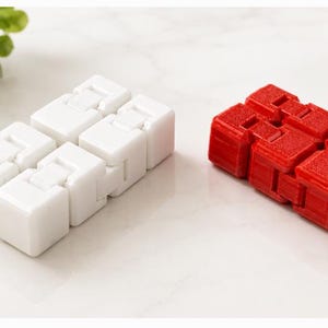 May include: Two fidget toys are displayed on a white surface. One is white, the other is red. Each is composed of connected, rectangular blocks. The white one is on the left, and the red one is on the right. Green leaves are visible in the background.