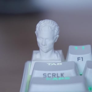 Puede incluir: Un teclado blanco con una tecla personalizada que presenta un busto. La tecla personalizada está colocada en la tecla Tab. Las teclas del teclado son blancas con letras y símbolos verdes iluminados.