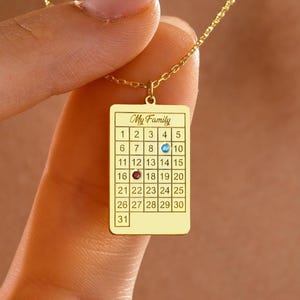 May include: A gold pendant necklace with a rectangular charm displaying a calendar design. The calendar is labelled "My Family" and has numbers 1-31. The necklace is held by a person.
