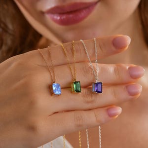 May include: A selection of gemstone pendant necklaces in gold and silver tones. Each necklace features a rectangular-cut gemstone in blue, green, or purple. The necklaces are displayed against a soft background.