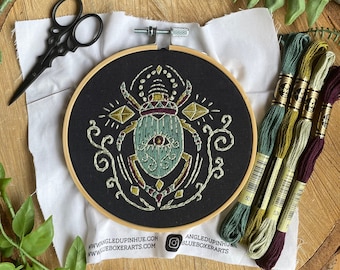 May include: A black embroidery hoop with a green and gold beetle design. The beetle has a large eye and is surrounded by geometric shapes. The hoop is on a wooden surface with green foliage and embroidery floss.
