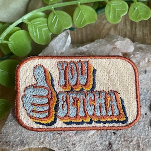 May include: A beige embroidered patch with a thumbs up and the text "You Betcha!" in a retro font with orange, yellow, and blue accents.