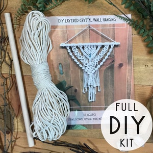 May include: A DIY layered crystal wall hanging kit. The kit includes string, a dowel, and wire. The image shows a completed white macrame wall hanging with a triangular design, a bundle of white string, and a wooden dowel. The words "FULL DIY KIT" are in a circle.