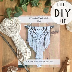 Op de afbeelding: Een doe-het-zelf macramé wandkleed kit, met een wit macramé ontwerp, katoenen koord en een houten stok. De kit bevat instructies voor het maken van een decoratieve wandhanger. De woorden "FULL DIY KIT" worden in een cirkel weergegeven.