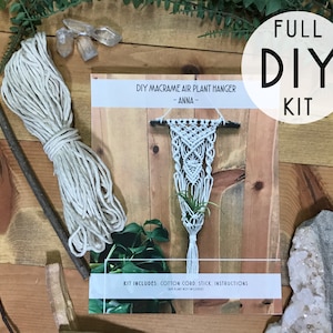 May include: A DIY macrame air plant hanger kit, featuring a white macrame plant holder, cotton cord, a wooden stick, and instructions. The kit includes the text "FULL DIY KIT" in a white circle. The kit is displayed on a wooden surface.