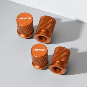 Puede incluir: Cuatro tapones de válvula de neumático de color naranja con superficie texturizada. Cada tapón tiene el texto "MX-5" en blanco en la parte superior. Los tapones están diseñados para atornillarse a una válvula de neumático.