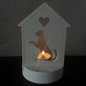 May include: A white wooden candle holder shaped like a house, featuring a heart cutout and a dog silhouette. A lit candle inside casts a warm glow. The holder has a small, rounded base.