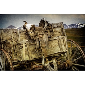 May include: An antique wooden wagon with a weathered appearance, featuring a brown leather saddle. The wagon is set against a backdrop of mountains and a cloudy sky, evoking a sense of the American West.