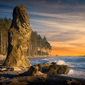 May include: A scenic coastal landscape featuring a large, weathered rock formation on a beach. The sky transitions from blue to orange, with a line of trees in the background. Waves crash on the shore, creating a dynamic scene.