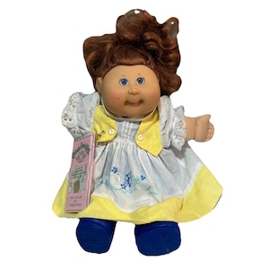 May include: A Cabbage Patch Kids doll with brown hair and blue eyes. The doll wears a white dress with blue floral embroidery, a yellow vest, and blue shoes. A pink tag is attached to the doll.