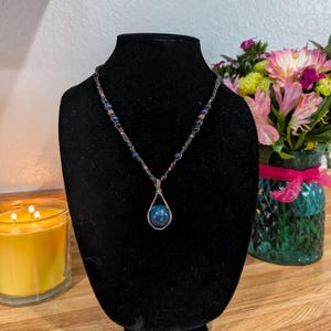 May include: A necklace featuring a dark wire-wrapped teardrop pendant with a round blue stone. The chain is composed of dark metal links interspersed with small blue beads and metallic accents, displayed on a black velvet-covered bust form.
