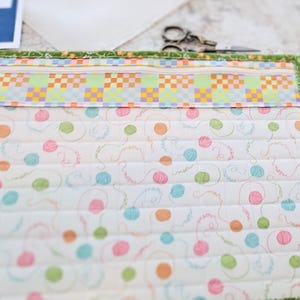 May include: A quilted pouch with a zip closure. The top section has a colourful patchwork design, while the main body displays a pattern of pastel-coloured yarn balls on a white background. The edges are trimmed with green floral fabric.