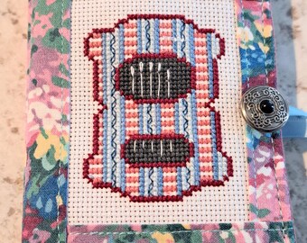 Puede incluir: Un estuche de agujas rectangular de tela con un estampado floral en tonos rosa, azul y amarillo. El panel central muestra un diseño de punto de cruz de un objeto a rayas con dos secciones gris oscuro que contienen agujas blancas, asegurado con un botón plateado.