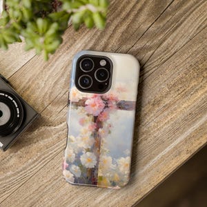 May include: A phone case featuring a cross adorned with pink and white flowers against a soft blue background. The case has a glossy finish and is set on a wooden surface, with a camera and greenery in the background.