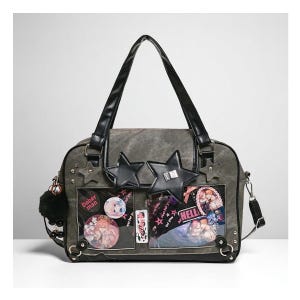 Peut inclure: Sac fourre-tout gris avec des poignées noires et une bandoulière amovible. Le sac présente des ornements en forme d'étoile, des épingles décoratives et des poches transparentes affichant des graphiques et du texte colorés. Un pompon noir est suspendu sur le côté.