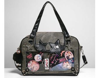 May include: Gray tote bag with black handles and a detachable shoulder strap. The bag features star-shaped embellishments, decorative pins, and clear pockets displaying colorful graphics and text. A black pom-pom hangs from the side.
