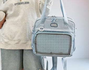May include: A light blue backpack-style tote bag with a clear mesh front pocket. The bag has two top handles and adjustable shoulder straps. The text "LUCKY FASHION" is on a patch on the front. The bag is held by a person wearing a gray hoodie.