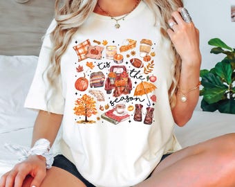 May include: A cream-colored t-shirt featuring a colorful autumn-themed graphic print with the text 'tis the season'. The design includes a red truck, pumpkins, a sweater, a football, an umbrella, boots, a tree, and various fall-related items.
