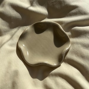 Op de afbeelding: Een kleine, beige keramische schaal met een golvende, organische vorm. De schaal is onversierd en ligt op een crèmekleurige stof, met schaduwen die diepte toevoegen. De handgemaakte schaal is waarschijnlijk bedoeld voor het vasthouden van kleine voorwerpen.