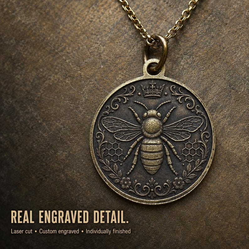 May include: A round bronze-toned pendant necklace featuring an embossed bee at the center, flanked by honeycomb patterns and floral motifs, topped with a small crown. The text REAL ENGRAVED DETAIL. Laser cut &bull; Custom engraved &bull; Individually finished.