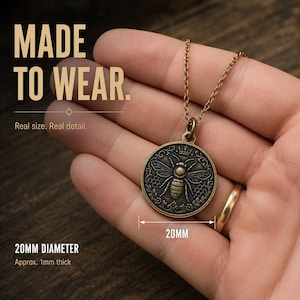 May include: A bronze-toned circular pendant necklace featuring an embossed bee design with a crown and honeycomb pattern. The pendant has a 20mm diameter and is approximately 1mm thick. Text on the image reads MADE TO WEAR. Real size. Real detail. 20MM DIAMETER Approx. 1mm thick.