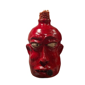 May include: A glossy red ceramic bottle shaped like a human head with a cork stopper. The face features white eyes with black pupils, a prominent nose, and a dark circular object held in the mouth. The surface has a smooth, reflective finish.