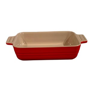 May include: A rectangular ceramic baking dish with a vibrant red exterior and a smooth cream-colored interior. The dish features two integrated side handles with a ribbed texture, providing a secure grip for moving the vessel.