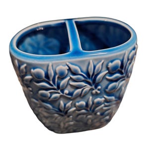 Puede incluir: Un porta cepillos de dientes de cer&aacute;mica azul con dos compartimentos. El soporte presenta un dise&ntilde;o floral en relieve en blanco y azul. El interior es de un tono azul m&aacute;s oscuro y el borde es azul m&aacute;s claro.