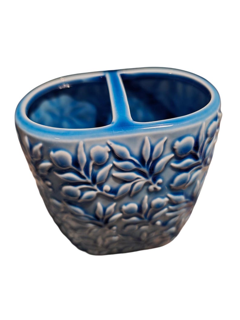 Puede incluir: Un porta cepillos de dientes de cer&aacute;mica azul con dos compartimentos. El soporte presenta un dise&ntilde;o floral en relieve en blanco y azul. El interior es de un tono azul m&aacute;s oscuro y el borde es azul m&aacute;s claro.