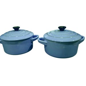 May include: Two light blue enameled cast iron round Dutch ovens with matching lids. Each pot features two side handles and a dark blue knob on the lid. The Le Creuset brand name is embossed on the top of each lid. The pots have a smooth finish.