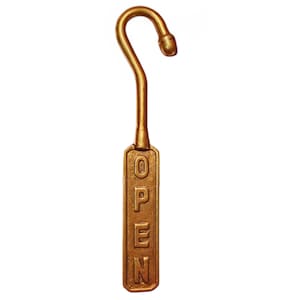 May include: A gold-toned metal sign featuring a vertical rectangular plaque with the word OPEN embossed in capital letters. The plaque is attached to a curved hook at the top, designed for hanging. The finish has a metallic, slightly textured appearance.