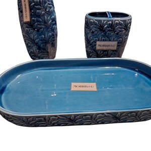 Puede incluir: Un juego de ba&ntilde;o de cer&aacute;mica azul que incluye un jabonera, un porta cepillos de dientes y un jarr&oacute;n alto. El juego tiene un patr&oacute;n de hojas y el texto "MORRIS & CO" en cada pieza. La jabonera es ovalada.