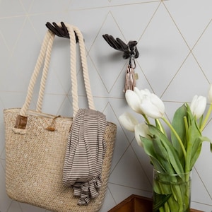 May include: Two dark brown wall-mounted hooks shaped like human hands. One hook supports a beige woven tote bag with thick rope handles and a striped fabric accessory, while the other hook holds a silver keychain with a metallic tassel charm.