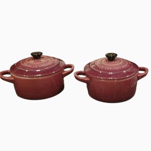 May include: Two round, plum-colored Le Creuset stoneware mini cocottes with matching lids. Each pot features two side handles, a dark knob on the lid, and the embossed Le Creuset logo on the lid surface. The finish is a glossy, deep purple.