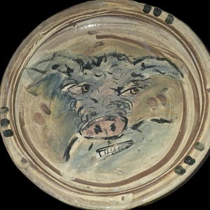 May include: A ceramic plate with a hand-painted pig face design. The pig's face is painted in shades of blue, pink, and brown, with a grumpy expression. The plate has a light brown rim with dark brown and black accents.