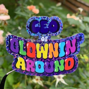 May include: A colorful sticker with the text "CEO OF BLOWIN' AROUND" in bubble letters. The sticker has a glittery border and is held by tweezers against a blurred background of green foliage and flowers.
