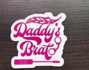May include: A white sticker with the words "Daddy's Brat" in a pink, cursive font. The sticker features a feather, handcuffs, and a paddle design. The sticker is on a dark brown wooden surface.