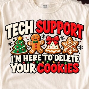 May include: A cream-coloured t-shirt featuring a graphic with the text TECH SUPPORT I'M HERE TO DELETE YOUR COOKIES. The design includes a gingerbread tree, a gingerbread person, a decorated bell, and a snowflake cookie with small lights.
