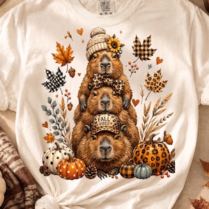 以下が含まれることがあります： クリーム色のTシャツで、3匹のカピバラが積み重なったグラフィックが描かれています。一番上のカピバラはヒマワリ付きのニット帽、真ん中はヒョウ柄の蝶ネクタイ、一番下はヒョウ柄のキャップを被っており、キャップにはFALL VIBESという文字が記されています。