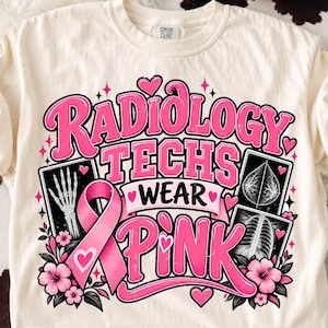 Op de afbeelding: Een crèmekleurig t-shirt met een opdruk met de tekst "RADIOLOGY TECHS WEAR PINK" in dikke roze letters. Het ontwerp bevat een roze bewustwordingslint, röntgenfoto's van een hand en borstkas, en roze hibiscusbloemen als accenten.