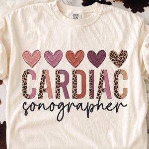 May include: A cream-colored t-shirt featuring the text CARDIAC in large block letters with a mix of solid pink, mauve, and leopard print patterns. Above the text are five hearts in similar colors and patterns, with the word sonographer below.