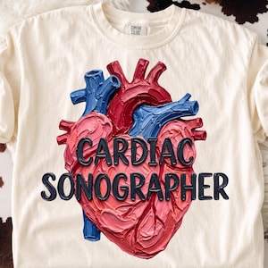 May include: A cream-colored short-sleeve t-shirt featuring a central graphic of an anatomical heart rendered in a textured, impasto-style paint effect with red and blue tones. The black text CARDIAC SONOGRAPHER is printed across the heart.