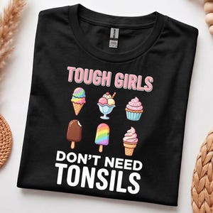 May include: A black t-shirt featuring the text TOUGH GIRLS DON'T NEED TONSILS in white and pink lettering. The design includes six colourful illustrations of various ice cream treats, including cones, a sundae, a cupcake, and a popsicle.