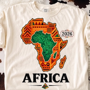 Könnte beinhalten: Ein cremefarbenes Kurzarm-T-Shirt mit einer Grafik des afrikanischen Kontinents. Die Karte ist mit orangefarbenen, gelben und grünen geometrischen Mustern gefüllt. Rechts steht der Text 2026 und darunter ist das Wort AFRICA in Schwarz gedruckt.