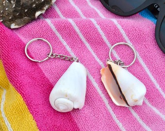 May include: Two seashell keyrings with silver key rings and chains rest on a pink, white, yellow, and blue striped towel. One keyring features a white conch shell, and the other has a shell with brown accents. Black sunglasses are also visible.