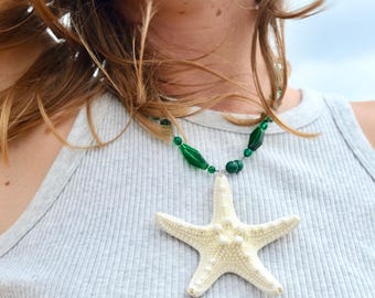 May include: A necklace featuring a large, off-white starfish pendant. The necklace has a strand of green beads and a silver-colored clasp. The starfish pendant is the focal point, with a textured surface and five prominent arms.