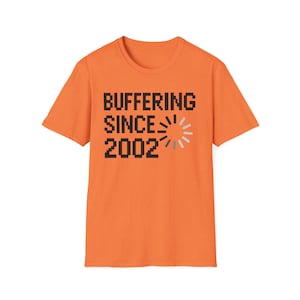 May include: Orange t-shirt with the text "BUFFERING SINCE 2002" in black pixelated font. A loading symbol is next to the text. The shirt is a solid orange color.