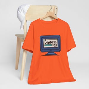 May include: Bright orange t-shirt with a blue computer monitor graphic displaying the word "LOADING". The screen also shows a loading bar and a cursor. A white baseball cap is on a wooden chair.
